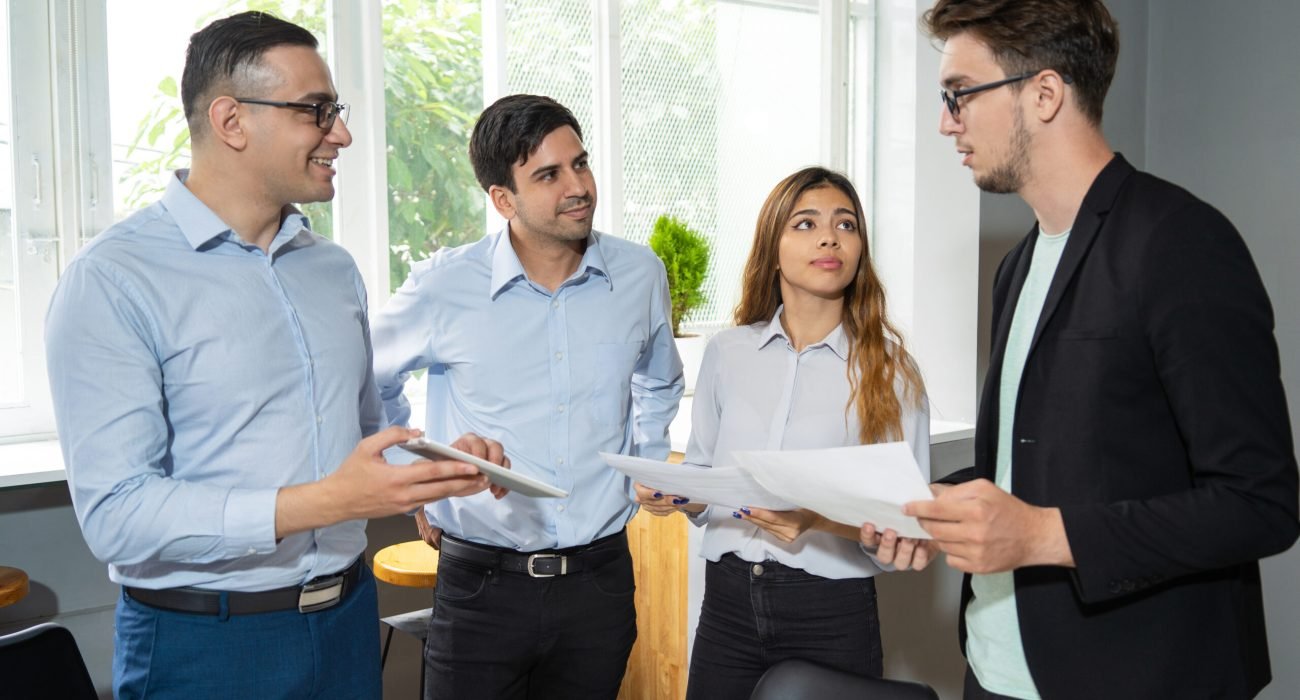 Workgroup of three reporting to serious young team leader. Coworkers showing tablet screen and papers to manager in jacket. Business team concept